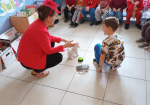 Nauczycielka prowadzi tematyczne zajęcia. W tle na ławeczkach siedzą dzieci.