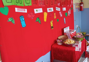 Na zdjęciu znajduje się tematyczna dekoracja, która wisi na tablicy. Na podłodze ułożone są pudła na zużyte baterie. Ponadto na stolikach widać przygotowane kosze ze słodyczami dla dzieci.