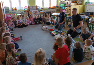 Dzieci siedzą w kole i słuchają wykładu prowadzonego przez Strażaków.