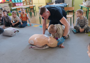 Strażak wspólnie z chłopcem prezentuje jak wykonać resustytację krążeniowo-oddechową.