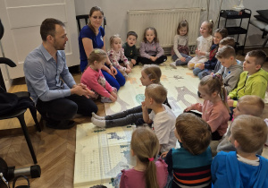 Dzieci siedzą na macie i słuchają wykładu prowadzonego przez fizjoterapeutę.