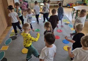 Dzieci uczestniczą w zabawie ruchowej. Na dywanie znajdują się kolorowe dyski.