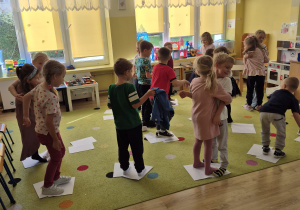 Dzieci bawią się na dywanie. Stoją obok siebie na kartkach papieru.