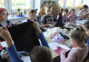grupa dzieci siedzi przy stoliku, na którym leży dużo różnych tkanin, wybierają, tną