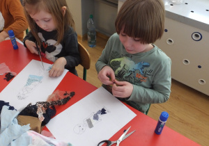 dwoje dzieci przy stoliku nakleja pocięte tkaniny na obrazek postaci